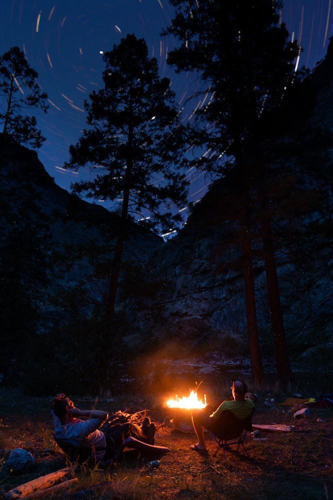 Riverside Camp. After a day on the river, two rafters enjoy the warmth of a fire and the spinning of the universe.